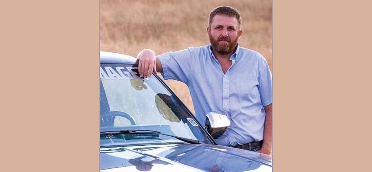 Independence Party of Alberta candidate Corrie Toone. White man with short reddish-brown hair, moustache and beard, leaning against a blue truck