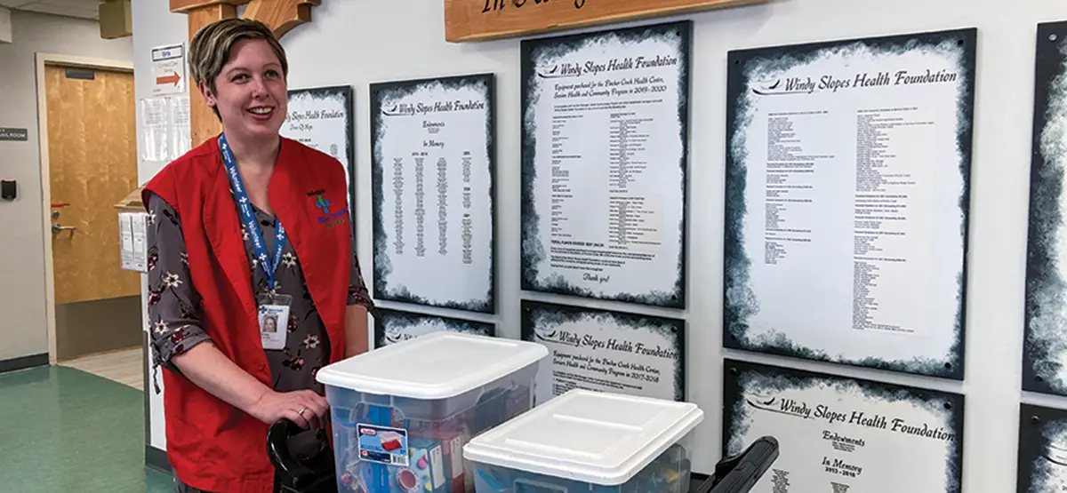 Rachel VanHalen, a woman with short brown hair and wearing a red vest, pushes a cart with activity bins at Pincher Creek Health Centre