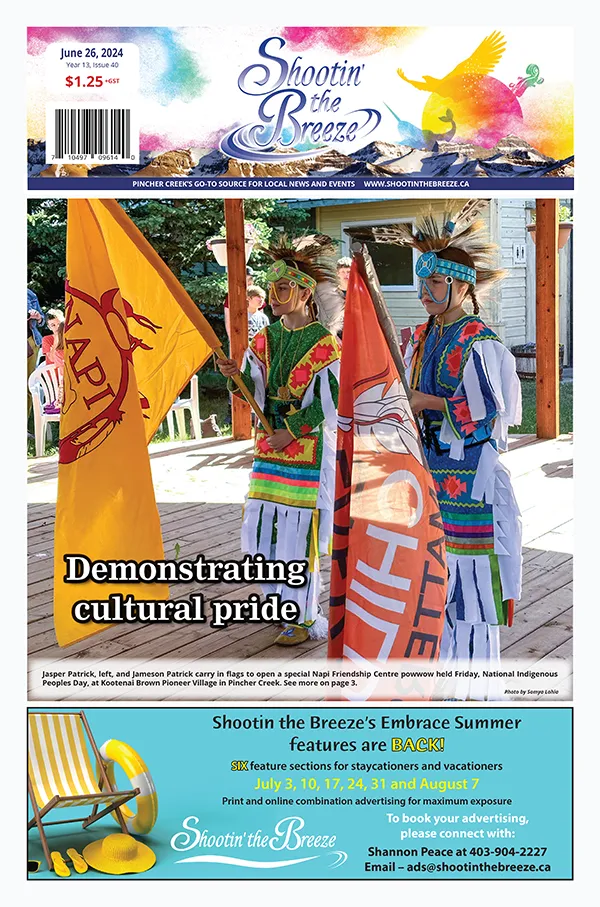 Front page of June 26, 2024, issue of Shootin' the Breeze — Jasper and Jameson Patrick, dressed in Indigenous grass dance regalia, carry a yellow flage with red Napi Friendship Association logo and orange Every Child Matters flag, open Indigenous Peoples Day powwow at Kootenai Brown Pioneer Village in Pincher Creek.