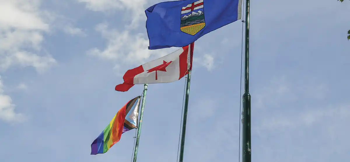 Three flags on poles waving in the wind against a blue sky: the Pride flag, the Canadian flag, and the provincial flag of Alberta.