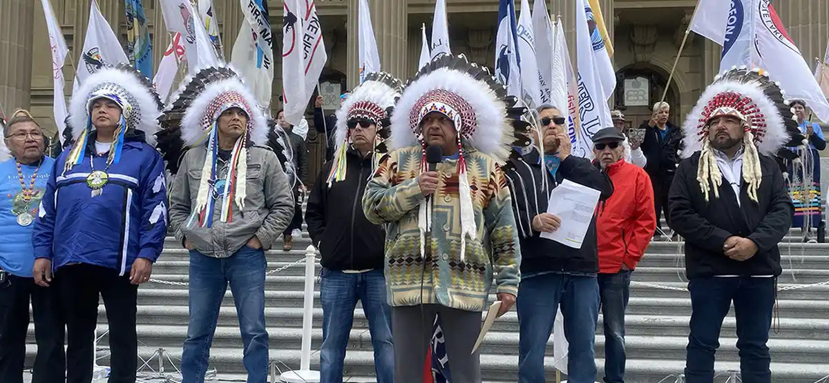 A group of Indigenous leaders, wearing traditional headdresses and regalia, stand together on steps with flags behind them. One leader speaks into a microphone while others stand in solidarity.