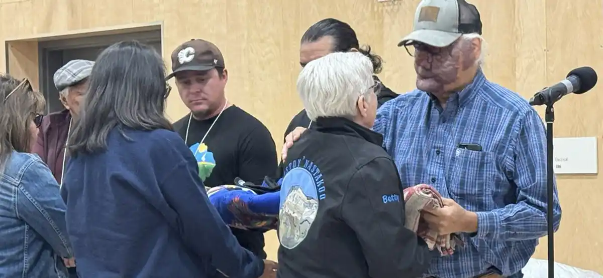 A group of people gather indoors as an older man in a cap and plaid shirt receives a folded blanket and a gift, while others stand nearby, some wearing jackets with mountain designs. A microphone is visible to the right.