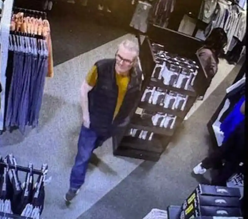 A man with short gray hair, glasses, a yellow shirt, a dark vest, and blue jeans walks through a clothing store, passing racks of apparel and shelves of packaged socks.