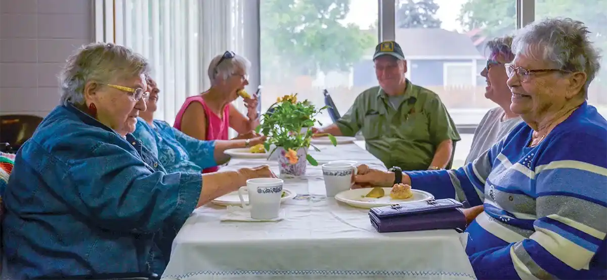 A group of older adults sit around a table by a window, enjoying food and coffee together. The table is decorated with a vase of flowers, and everyone appears to be talking and smiling in a bright, cheerful room.