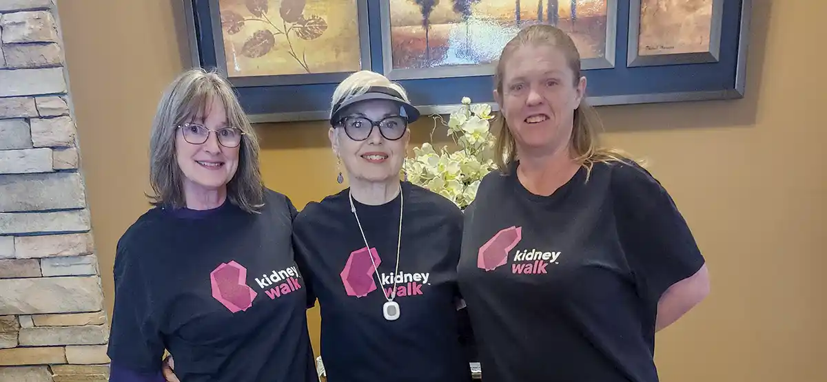 Three women stand side by side indoors, smiling and wearing matching black t-shirts that read “kidney walk” with a pink logo. There are framed pictures and a vase with white flowers in the background.