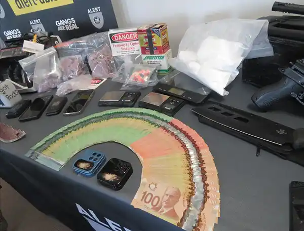 A table displays stacks of Canadian $100 bills, several mobile phones, bags of white powder, packages, a handgun, and various other items, suggesting evidence from a police investigation or seizure.