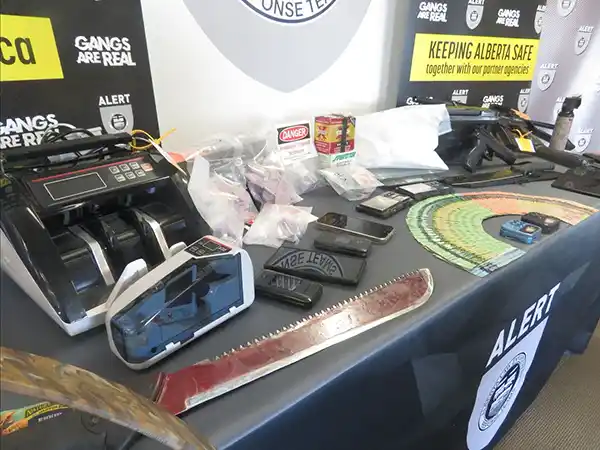 A table displays evidence including a machete, money counter, scales, packaged items, a taser, and various tools. ALERT and “Gangs Are Real” banners are visible in the background, indicating a law enforcement presentation.