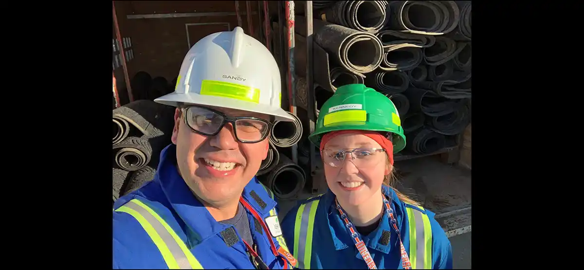 Two smiling workers wearing blue coveralls, safety vests, and hard hats (white and green) pose for a selfie in front of stacked rolled-up mats or industrial materials at a worksite.