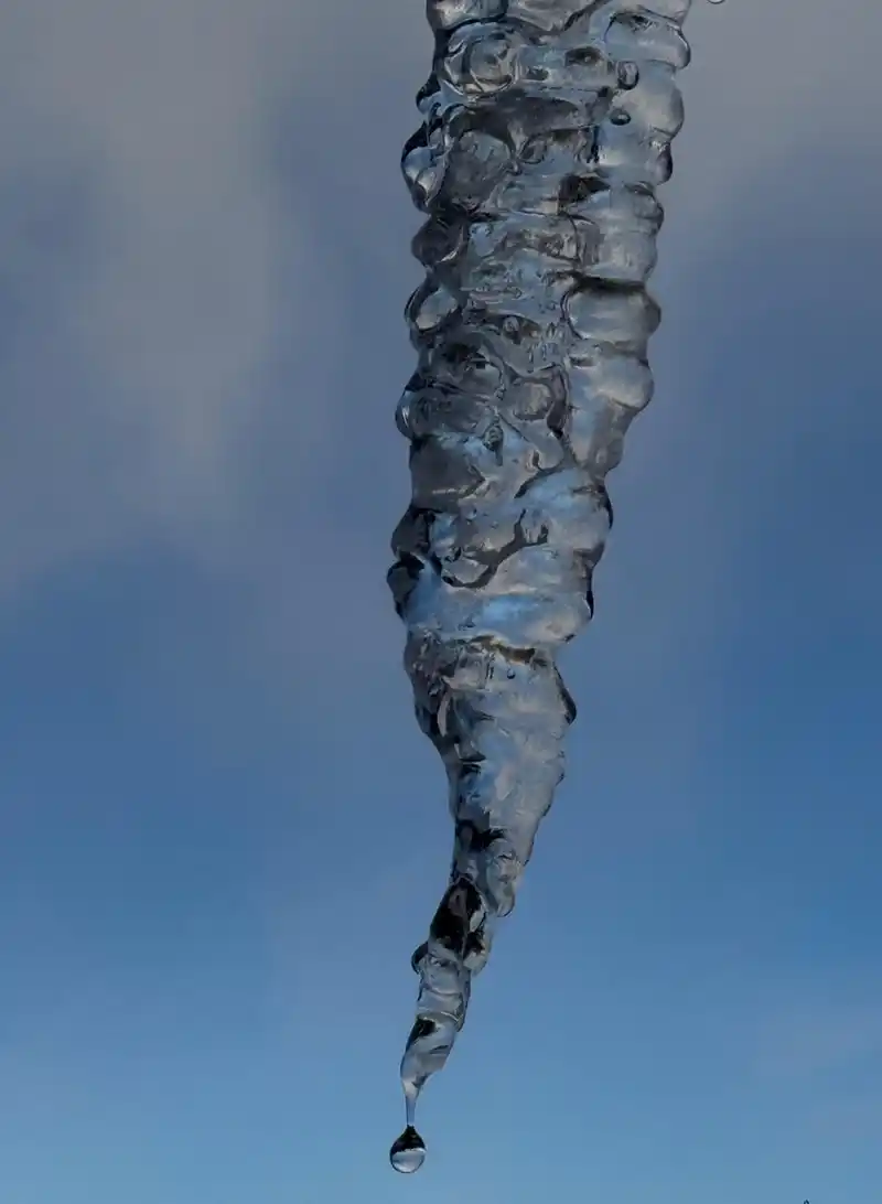 A close-up of a clear, textured icicle with a droplet of water forming at its pointed tip, set against a blue sky with faint clouds in the background.