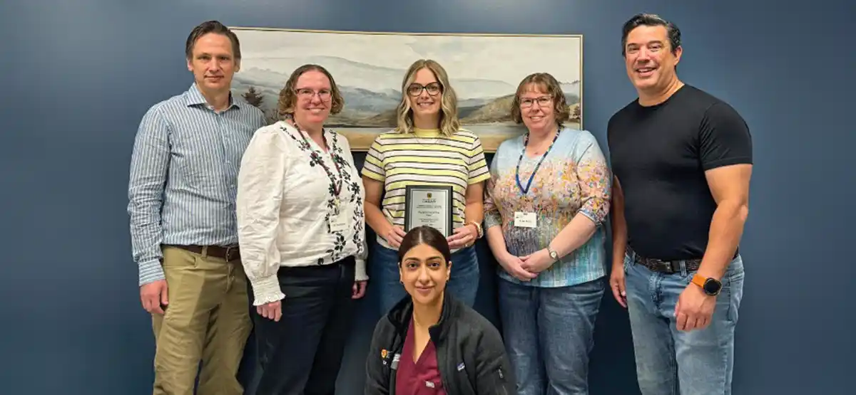 Six people pose indoors, one kneeling in front. The center person holds a framed certificate. They stand against a blue wall with a landscape painting behind them. All are smiling.